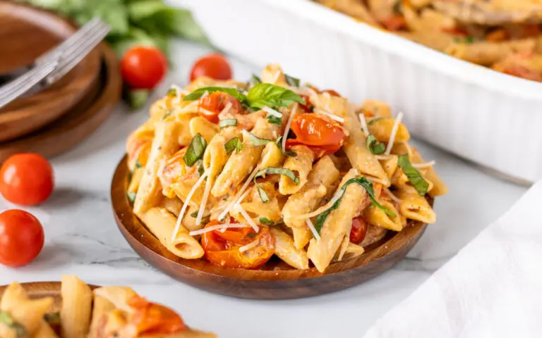 Cheesy baked pasta with tomatoes in a serving bowl with a wooden bowl and tomatoes behind the plate of pasta.