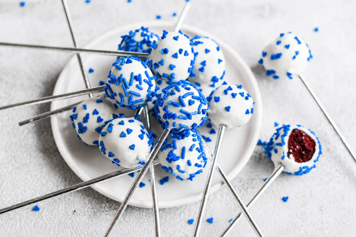 White chocolate cake pops covered in blue sprinkles on metal sticks arranged on a white plate, with one bite showing a red center inside the pop.