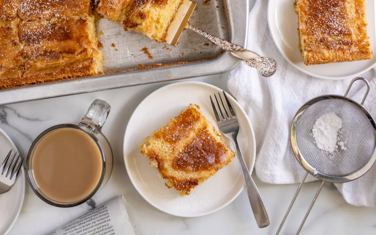 Ooey Gooey Butter Cake on a plate with a fork and powdered sugar sprinkled around. A cup of coffee and pan of Ooey Gooey Butter Cake in the background.
