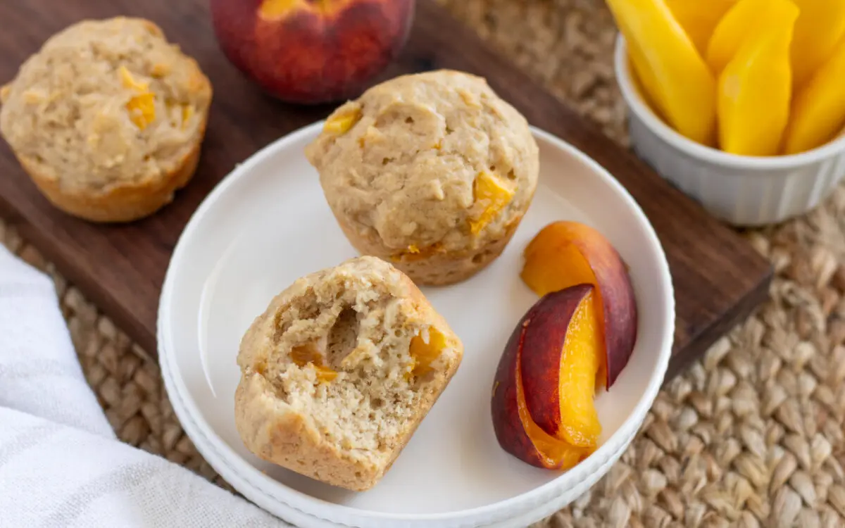 Fruit and Yogurt Muffins cut in half on a plate with peaches sitting on top of a wooden board and a dish cloth.