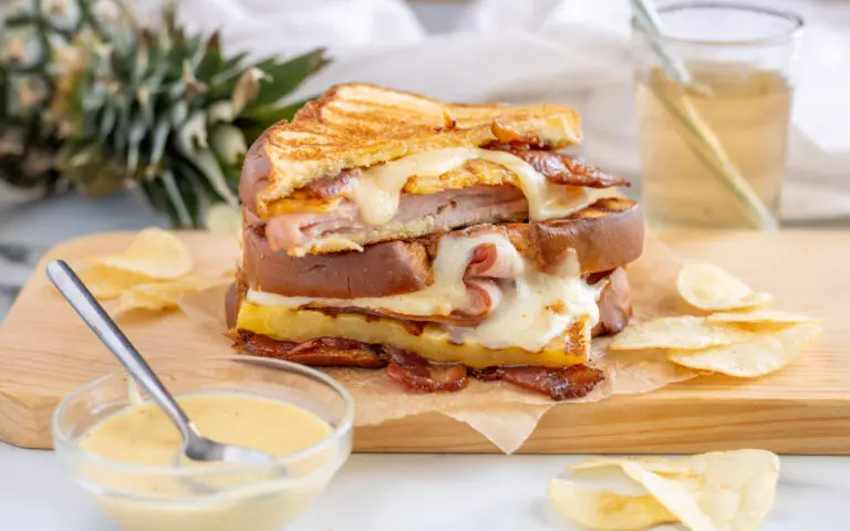Hawaiian Panini on top of a serving platter cut in half with chips around the serving platter. Hot Honey Mustard Aioli, a drink and pineapple are featured in the background.