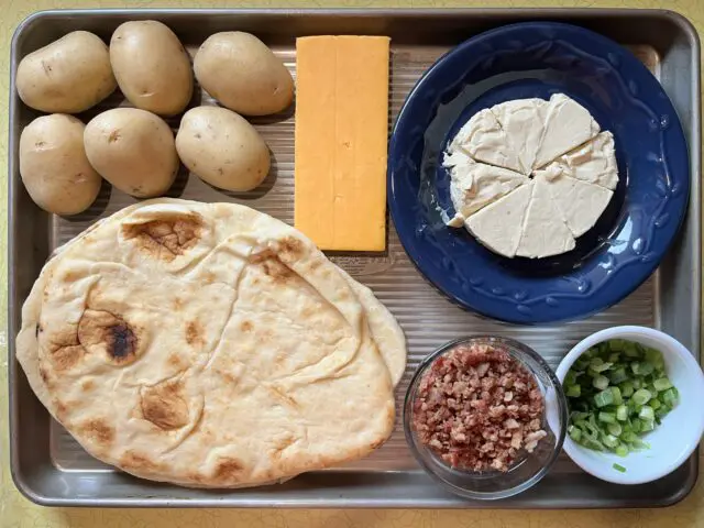 Loaded Potato Naan Pizza ingredients laid out. This pizza is made with naan flatbread, bacon bits, yellow potatoes, garlic and herb cheese spread, shredded cheddar cheese and green onions.