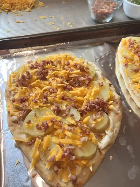 Loaded Potato Naan Pizza on a sheet pan getting ready to be baked after the potatoes and bacon bits are put on top of the pizza and the remainder of the cheese is sprinkled on top. 