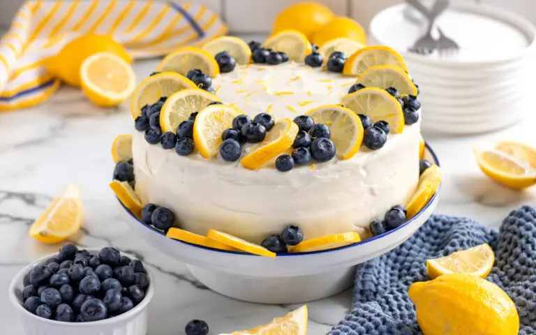 Lemon Blueberry Cake decorated with Lemons and Blueberries sitting on a cake stand. In the background there is blueberries, lemons and forks. Featuring vibrant blue and yellows from the citrus and fruit.