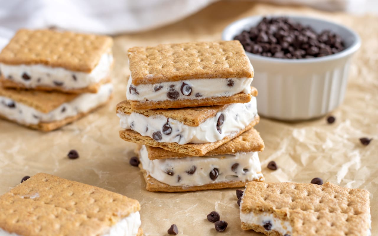 Three ice cream sandwiches stacked on top of each other with four other sandwiches scattered around in the background and a bowl of chocolate chips behind the stack of ice cream sandwiches.