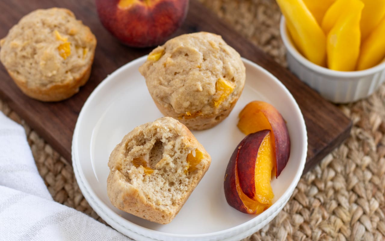 Fruit and Yogurt Muffins cut in half on a plate with peaches sitting on top of a wooden board and a dish cloth.